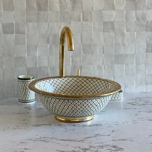 May include: A white ceramic sink with a black and gold geometric pattern. The sink has a gold rim and a gold base. The sink is sitting on a white countertop.