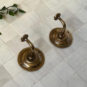 May include: Two antique brass wall hooks with a decorative design. The hooks are mounted on a white tiled wall.