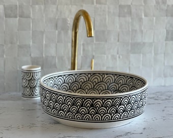 May include: A white ceramic vessel sink with a black and white geometric pattern. A matching tumbler is to the left. A gold faucet is in the background. The sink is a decorative bathroom fixture.