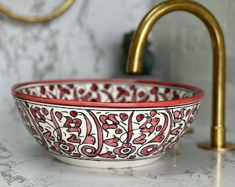 May include: A decorative ceramic sink with a floral design in red and black on a white background. The sink has a red rim and is next to a gold faucet. The sink is a unique bathroom fixture.
