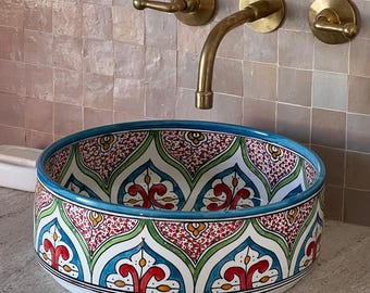 May include: A white ceramic sink with a colourful Moroccan-inspired design. The sink has a round shape and features intricate patterns in shades of red, green, blue, and yellow. The sink is set against a backdrop of pink mosaic tiles.