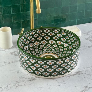 Op de afbeelding: Een witte keramische wastafel met een groen-wit bloemenpatroon. De wastafel is rond en heeft een lichte rand rond de rand. De wastafel staat op een wit aanrecht.
