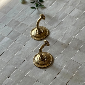 May include: Two gold-toned metal hooks with decorative details. The hooks are mounted on a white tiled surface.