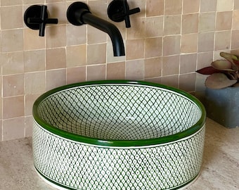 May include: Oval ceramic sink with a green and white geometric pattern. The sink has a green border and is paired with black fixtures. The sink is on a light-coloured countertop, with a tiled wall in the background.