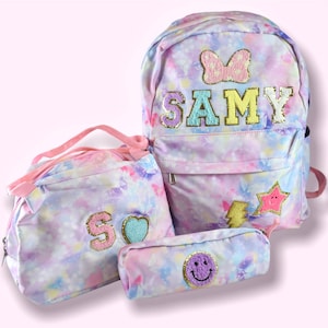 May include: A set of three matching school bags: a backpack, a lunch bag, and a pencil case. The bags feature a pastel watercolor design with butterflies and the name "SAMY" on the backpack. The pencil case has a smiley face.