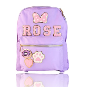 May include: A lavender purple backpack with a gold zipper and a variety of pink and gold patches. The patches include a bow, the name "ROSE", a donut, a paw print, and a heart.