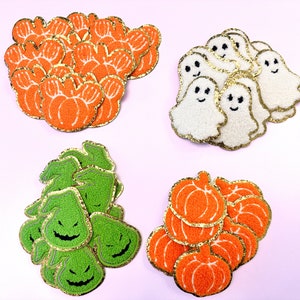 May include: A collection of Halloween-themed felt patches. There are orange pumpkins with gold glitter trim, white ghosts with gold glitter trim, and green jack-o'-lantern faces with gold glitter trim.