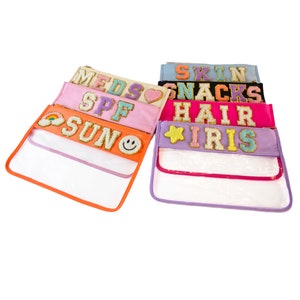 May include: A set of eight clear zippered pouches with colorful fabric fronts. Each pouch has a different word or phrase on the front, including "MEDS", "SPF", "SUN", "SKIN", "SNACKS", "HAIR", and "IRIS".