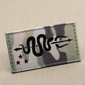 May include: A rectangular patch with a camouflage pattern. The patch features a black design of a snake intertwined with a trident and three stars. The patch is likely for military or tactical use.