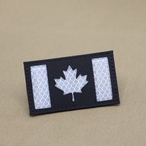 Könnte beinhalten: Ein schwarz-weißes kanadisches Flaggen-Patch mit reflektierendem Material. Die Flagge besteht aus schwarzem Stoff mit einem weißen kanadischen Ahornblatt und zwei weißen Streifen.