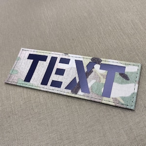 May include: A rectangular patch featuring the word "TEXT" in dark blue block letters. The patch has a camouflage pattern in green, brown, and tan. The patch is edged with a black border.