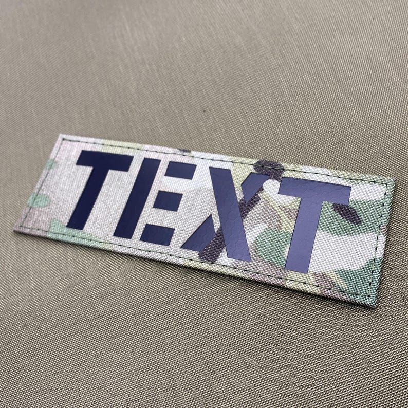 Pode incluir: Um patch retangular com a palavra "TEXTO" em letras mai&uacute;sculas azul escuro. O patch tem um padr&atilde;o de camuflagem em tons de verde, castanho e bege. O patch &eacute; contornado por uma borda preta.