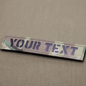 Può includere: Una toppa rettangolare con le parole "YOUR TEXT" in lettere nere. La toppa presenta un motivo mimetico nei toni del verde, marrone e beige. Lo sfondo è di colore beige.