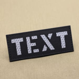 以下が含まれることがあります： 大きな反射性の白い文字で「TEXT」という単語が書かれた黒い長方形のパッチ。文字はハニカム模様です。パッチはベージュ色の背景にあります。