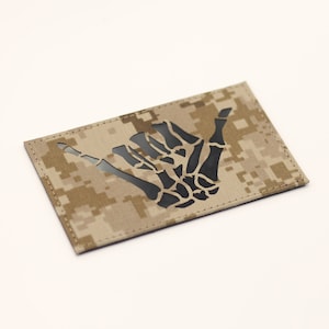 May include: A tan and brown camouflage fabric patch with a black skeleton hand making the shaka sign.