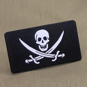 Könnte beinhalten: Schwarzer rechteckiger Aufnäher mit einem weißen Totenkopf- und gekreuzten Knochen-Design. Zwei weiße gekreuzte Schwerter befinden sich unter dem Totenkopf. Der Aufnäher hat eine strukturierte Oberfläche und einen schwarzen Rand. Das Design ist eine klassische Piratenflagge.