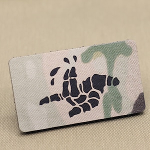 May include: Rectangular patch with a camouflage pattern in shades of green, tan, and gray. A black skeletal hand design is centered on the patch. The patch has a textured surface and a stitched border.