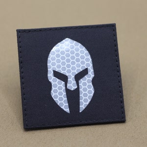 Op de afbeelding: Een zwarte patch met een wit reflecterend Spartaans helmdesign. De helm is gemaakt van een honingraatpatroon.