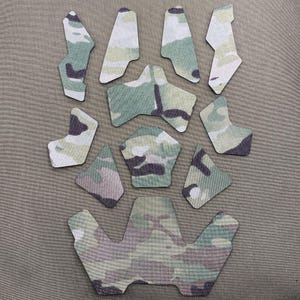 May include: A collection of camouflage-patterned helmet pads in various shapes and sizes. The pads feature a mix of green, brown, and white hues, designed for tactical or outdoor use. The pads are arranged on a neutral-colored surface.