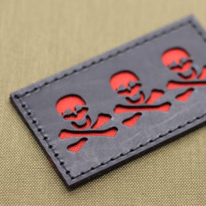 May include: A rectangular black patch featuring a red skull and crossbones design. The patch has a stitched border and is set against a tan background. The skull and crossbones are cut out of the black material, revealing the red backing.