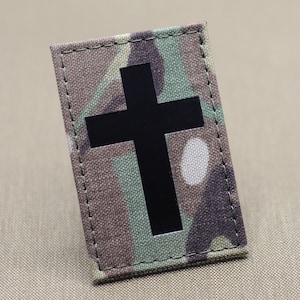 May include: A rectangular patch with a camouflage pattern in shades of green, brown, and tan. A solid black cross is centered on the patch. The edges are stitched with a matching thread.