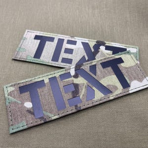 Pode incluir: Duas etiquetas retangulares com a palavra "TEXTO" em letras pretas. O fundo apresenta um padr&atilde;o de camuflagem em tons de verde, castanho e bege. As bordas das etiquetas s&atilde;o costuradas.