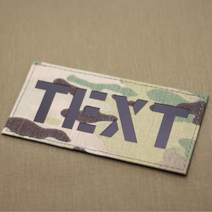 May include: A black patch with the word "TEXT" in white letters on a camouflage background.