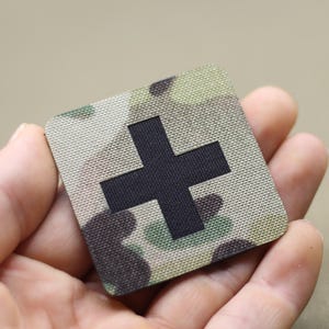 Könnte beinhalten: Ein quadratischer Patch mit einem schwarzen Pluszeichen auf einem Tarnmusterhintergrund. Der Patch ist aus einem gewebten Stoff.