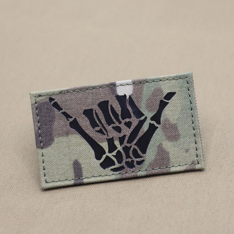 May include: A black skeleton hand making the shaka sign, a popular Hawaiian gesture, is embroidered on a camouflage fabric patch.