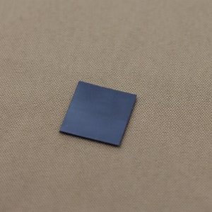 Op de afbeelding: Een klein, vierkant, donkerblauw object. Het object heeft een effen kleur en lijkt van een glad materiaal te zijn gemaakt. De achtergrond is een textiel van bruinachtige kleur. Het object staat in een lichte hoek.