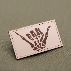 Könnte beinhalten: Ein weißer Leder-Patch mit einer braunen Skeletthand, die das Shaka-Zeichen macht. Der Patch ist an den Rändern vernäht.