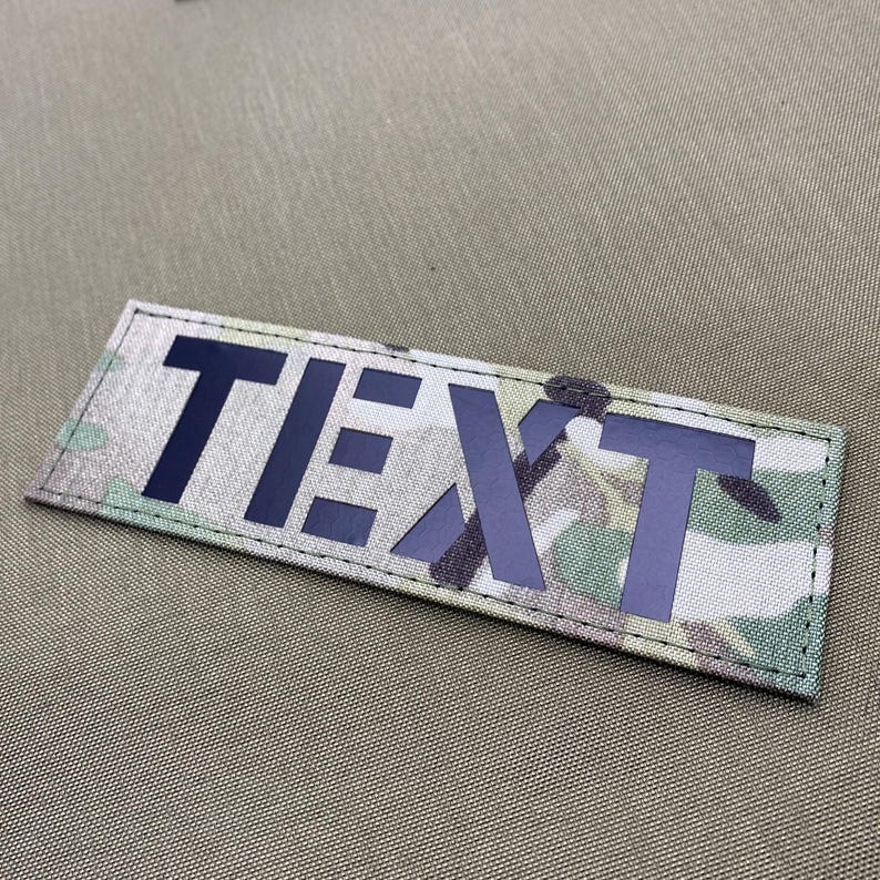 Pode incluir: Um patch retangular com a palavra "TEXT" em letras mai&uacute;sculas azul escuro. O fundo apresenta um padr&atilde;o de camuflagem em tons de verde, castanho e bege. O patch &eacute; costurado nas bordas.