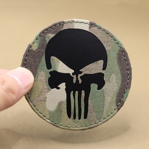 Pode incluir: Um remendo redondo de caveira preto e branco com fundo de camuflagem. A caveira é uma versão estilizada do logotipo do Punisher.