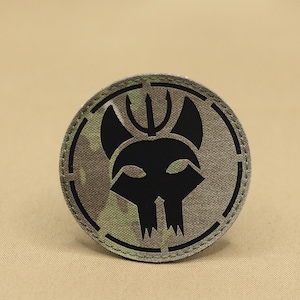 Può includere: Un cerotto mimetico nero e verde con una sagoma nera di un teschio con corna e un tridente sopra. Il cerotto è circolare e ha un bordo cucito.