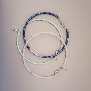 Può includere: Quattro braccialetti di perline nei toni del bianco, azzurro e blu navy, con chiusure e ciondoli argentati. I braccialetti sono disposti in un motivo circolare su uno sfondo grigio neutro. Le perline sono piccole e uniformi.
