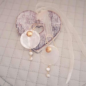May include: A pair of earrings with large, round, white shell pendants and gold details. Each earring has a small gold disc and three dangling white pearls. The earrings are presented on a wooden heart with a sheer ribbon, against a quilted background.