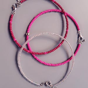 Pode incluir: Três pulseiras de contas em tons de rosa e transparente. As pulseiras são feitas de pequenas contas redondas enfiadas. Cada pulseira tem um fecho prateado. As pulseiras estão dispostas sobre uma superfície cinza.