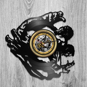May include: A black vinyl record clock with a gold rim and Roman numeral clock face. The clock is shaped like two figures reaching towards each other.