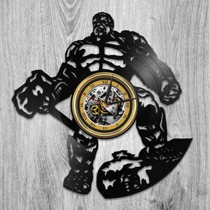 May include: Black wall clock in the shape of a muscular superhero. The clock face is gold with Roman numerals and visible gears. The superhero figure is cut from a black material, creating a silhouette.