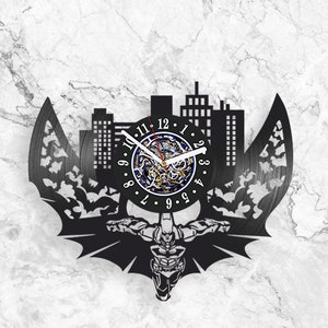Puede incluir: Reloj de vinilo negro con una silueta de Batman y una ciudad en el fondo. La esfera del reloj tiene un fondo negro con números blancos y un diseño azul y dorado en el centro.