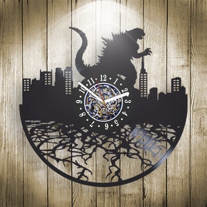 May include: Black vinyl record clock featuring a silhouette of Godzilla destroying a city. The clock face is white with black numbers and hands. The clock is mounted on a black background with a tree root design.