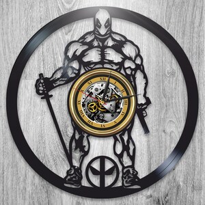 Puede incluir: Reloj de vinilo negro con una silueta de una figura musculosa que lleva una máscara y sostiene espadas. La esfera del reloj es dorada con números romanos y un mecanismo de reloj visible.