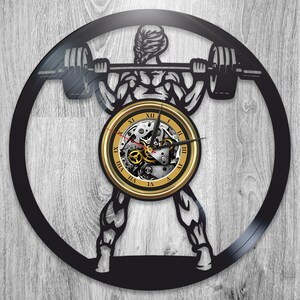 May include: Black vinyl record clock featuring a silhouette of a person lifting weights. The clock face is gold with Roman numerals and a visible clock mechanism.