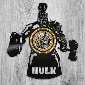 May include: Black vinyl record clock featuring a silhouette of the Hulk, a green superhero, holding a hammer. The clock face is gold with Roman numerals and a visible clock mechanism. The words "HULK" are printed in white below the clock face.