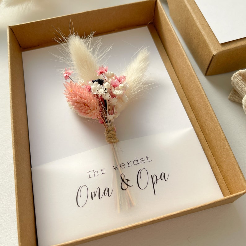 May include: A white card with a small bouquet of dried flowers in pink, white, and brown tones. The card is in a brown box and has the text "Ihr werdet Oma & Opa" written on it.