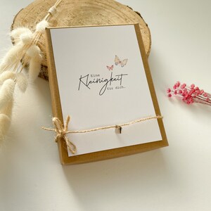 May include: A small brown box with a white card inside. The card has a handwritten message that says "Eine Kleinigkeit f&uuml;r dich." and two watercolor butterflies. The box is tied with twine.