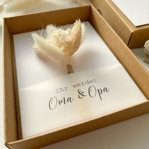 May include: A white card with the text "Ihr werdet Oma & Opa" in a brown box with a bouquet of dried white flowers on top.