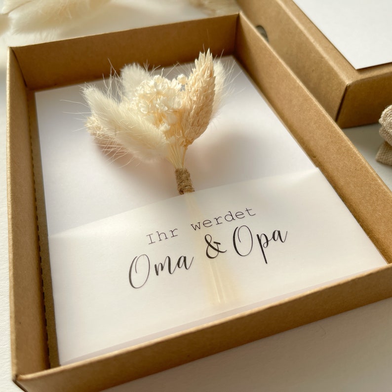 May include: A white card with the text "Ihr werdet Oma & Opa" in a brown box with a bouquet of dried white flowers on top.