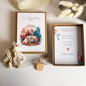 Könnte beinhalten: Eine Geburtstagskarte mit einer Aquarell-Illustration eines Wohnmobils in einer Waldlandschaft. Die Karte befindet sich in einem braunen Umschlag und trägt den Text "Zum Geburtstag Mara" auf der Vorderseite. Die Karte enthält außerdem eine kleine Geschenktüte, ein Glasröhrchen mit einem Korkstopfen und eine kleine Karte mit dem Text "Zum Geburtstag habe ich zwei gute Ratschläge für dich 1. Vergiss die Vergangenheit. Du kannst sie nicht ändern. 2. Vergiss dein Geschenk. Ich hab eins dabei. Paula & Robert".