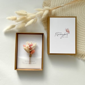 May include: A small, brown box with a white card inside. The card has a small bouquet of dried flowers and the text "Mochtest du meine Trauzeugin sein?" The box is closed and has a white card on top with the text "Eine Kleinigkeit fur dich." and a pink butterfly.